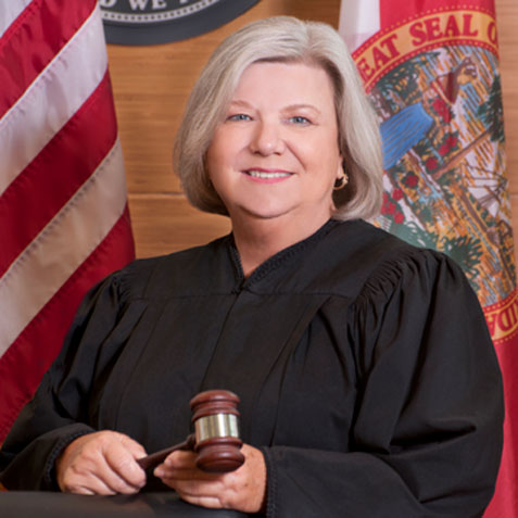 This is an image of a woman dressed in a black robe holding a gavel and standing in front of two flags.