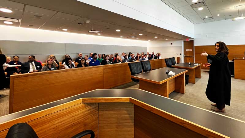 This is an image of a woman dressed in a black robe speaking to a group of people seated in the back of a courtroom.