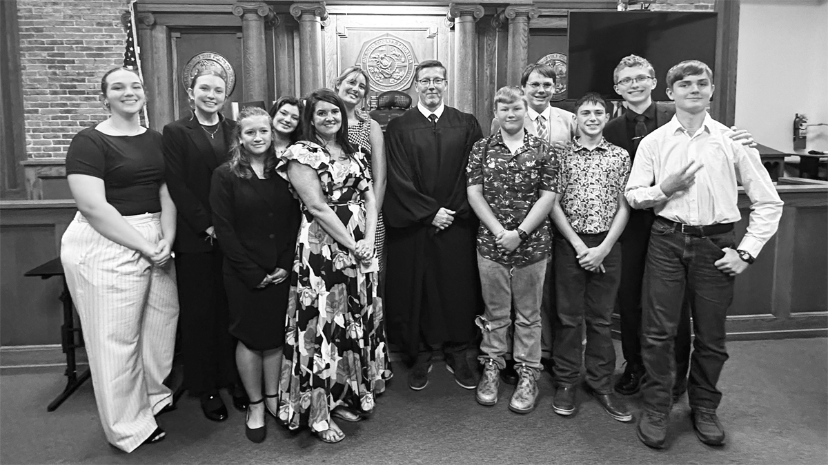 Eleven people, teacher and students, plus one judge stand in a courtroom. The judge is dressed in a black robe.